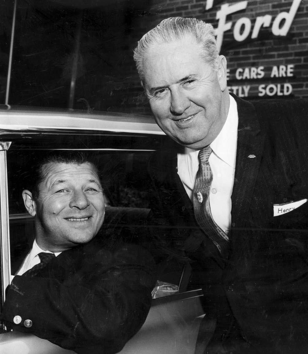Black and white photo of a smiling man in a car, another smiling man leaning in from outside. Ford sign in background.