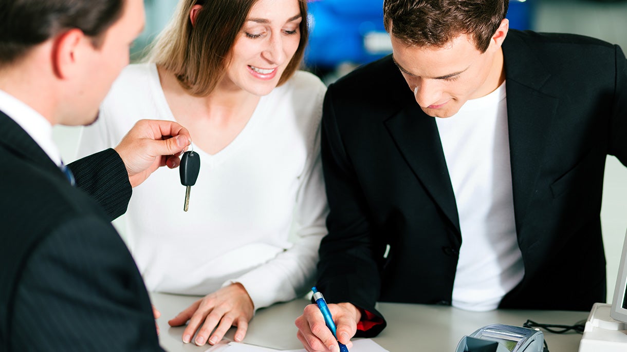 A salesperson handing car keys to a man signing paperwork next to a woman at a dealership counter.