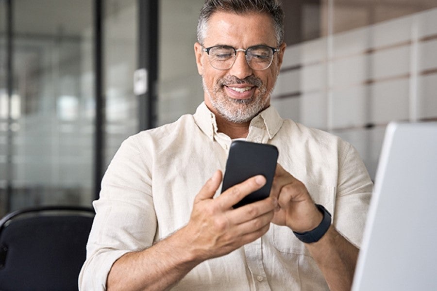 A smiling man with glasses and a beard using a smartphone in an office setting.