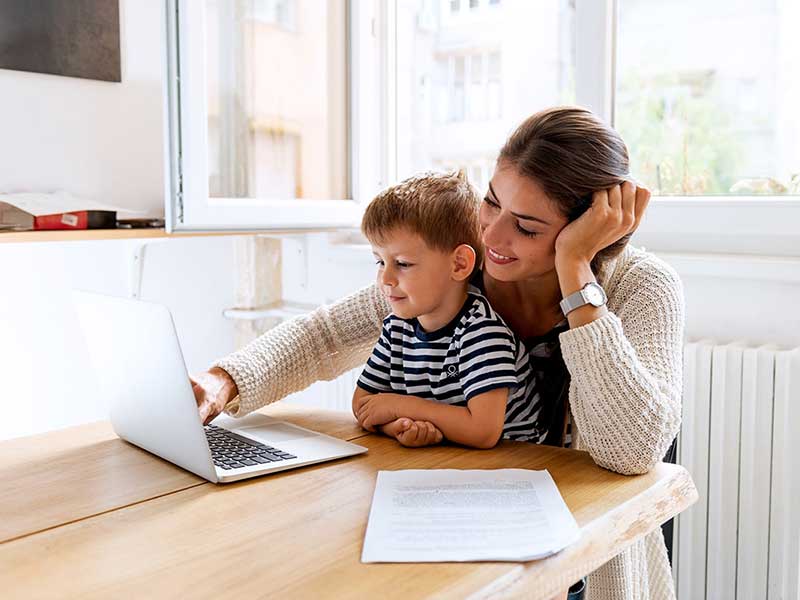 A smiling woman with brown hair and a young boy look at a laptop on a wooden table.